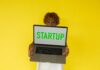 India’s AI Startups Dominate $10.1 Billion FY26 Funding Despite Overall Decline Startup laptop held by person against yellow background.